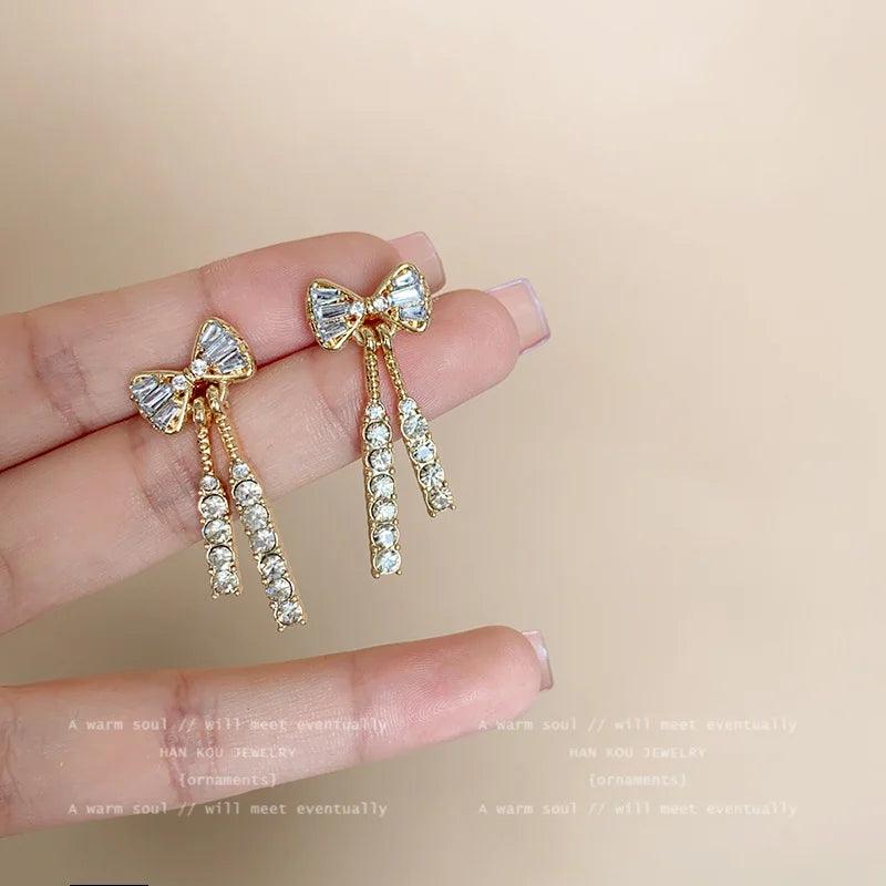 WOLZI Elodie Crystal Bow Stud Earrings with dangling rhinestone tassels held in hand